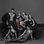 High-fashion editorial group portrait featuring six models in avant-garde black outfits with gold accents, dramatic poses, and regal accessories against a dark gray backdrop