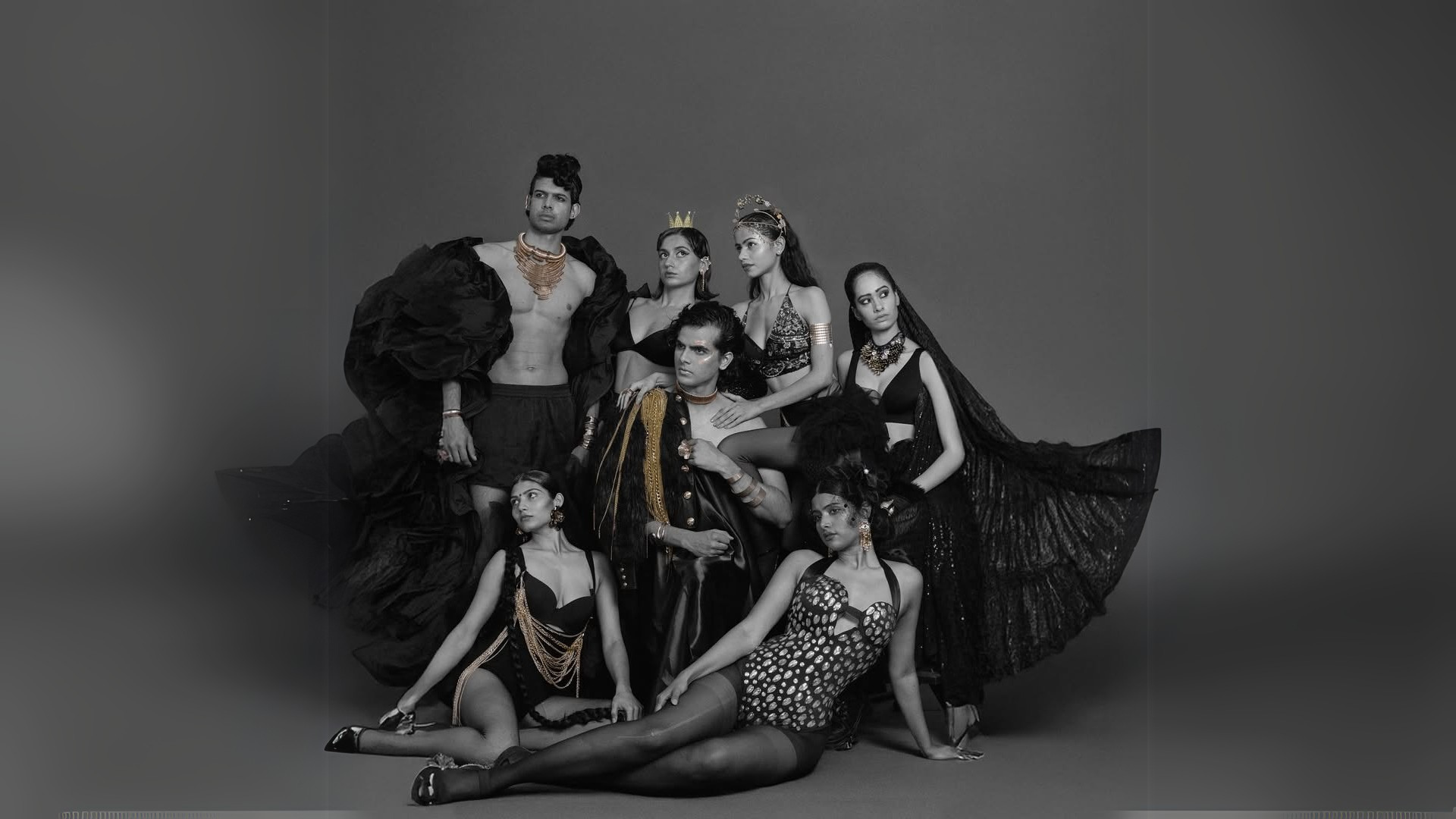 High-fashion editorial group portrait featuring six models in avant-garde black outfits with gold accents, dramatic poses, and regal accessories against a dark gray backdrop